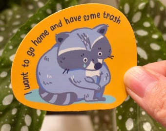 May include: A die-cut sticker featuring a sad cartoon raccoon in blue and gray, with the text "I want to go home and have some trash" on an orange background. The sticker is held in front of a green plant.