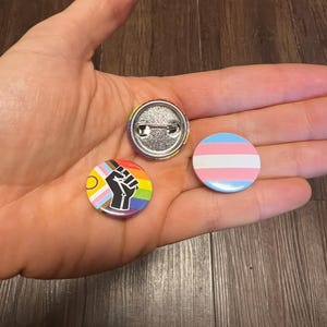 May include: Three circular pin badges with vibrant designs. One badge displays a rainbow and a raised fist, another features the transgender flag colours, and the third has a rainbow pattern. The badges are held in a hand.