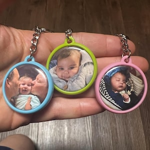 May include: Three round keychains with baby photos. Each keychain has a different colored border: blue, green, and pink. The keychains are attached to silver chains. The photos show babies.