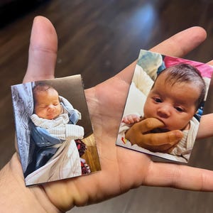 Op de afbeelding: Twee kleine rechthoekige foto's van een baby. Eén toont de baby in een blauwe deken gewikkeld, de andere toont de baby met een hand in de buurt van zijn mond. De baby heeft donker haar en een lichte huid. De foto's worden in een hand gehouden.