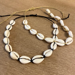 May include: Two cowrie shell necklaces. The shells are strung on black and tan cords. The necklaces are arranged on a wooden surface. The shells are white with a natural, smooth texture. The cords are knotted to adjust the length.