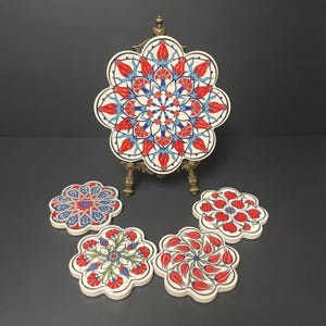 May include: A collection of decorative ceramic coasters with a floral design. The coasters are white with red, blue, and green patterns. One large coaster is displayed on a small brass easel. The coasters are approximately 4 inches in diameter.