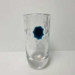 Pode incluir: Um vaso de vidro transparente com um design em forma de estrela azul escuro no centro. O vaso tem uma forma cilíndrica com uma base arredondada e um padrão de pequenas bolhas transparentes nas laterais.
