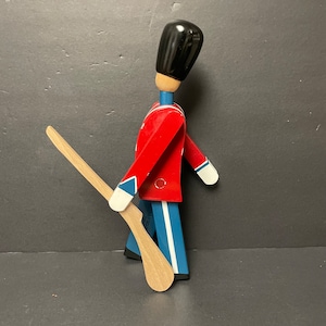 May include: A wooden toy soldier with a red coat, blue pants, and a black hat. The soldier is holding a wooden rifle.