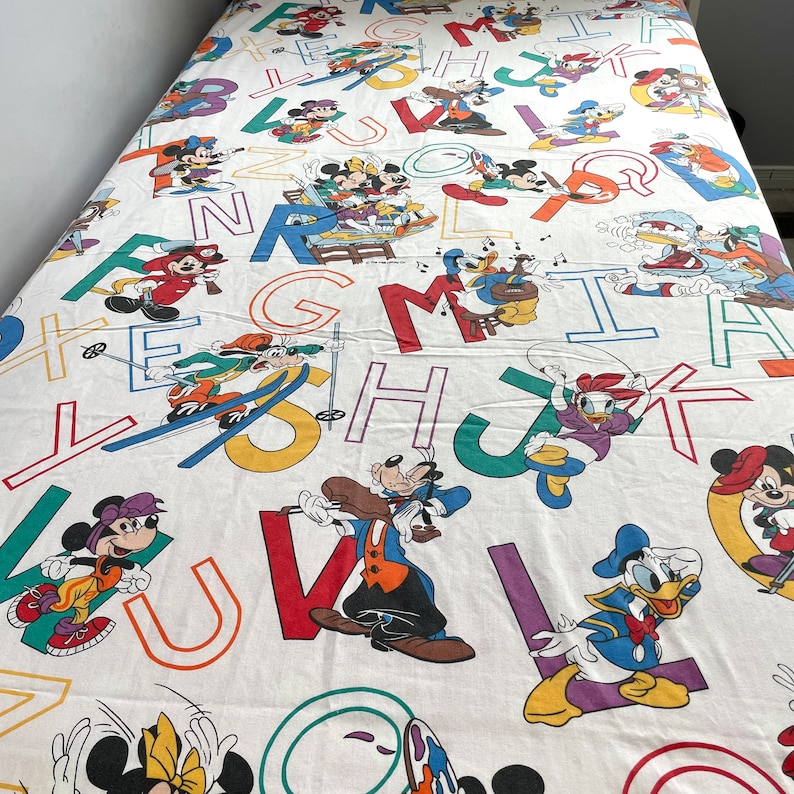 K&ouml;nnte beinhalten: Wei&szlig;es Bettlaken in Zwillingsgr&ouml;&szlig;e mit einem bunten Alphabetdruck mit Mickey Mouse, Minnie Mouse, Donald Duck, Goofy und Pluto. Die Figuren sind in verschiedenen Aktivit&auml;ten dargestellt, wie z. B. Musik spielen, Skifahren und Sport treiben.