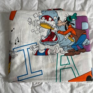 K&ouml;nnte beinhalten: Ein wei&szlig;er Baumwollstoff mit einem bunten Comic-Druck eines Charakters, der einem Flusspferd die Z&auml;hne putzt. Der Stoff hat die Buchstaben 'I' und 'A' aufgedruckt.