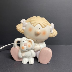 Puede incluir: Una figurita de cerámica de una niña con cabello rubio y un vestido rosa que sostiene un cachorro blanco. La niña tiene las mejillas sonrosadas y una expresión dulce. La figurita es una pieza de decoración encantadora y caprichosa.