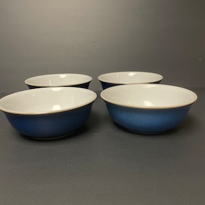 May include: Four ceramic bowls with a white interior and a blue exterior. The bowls have a rounded shape and a simple design. The bowls are arranged in a group on a dark surface.