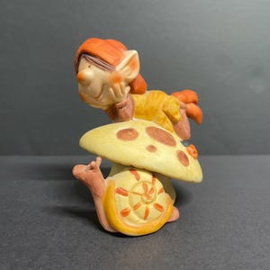 Puede incluir: Una caprichosa figura de cerámica que representa un elfo sonriente con cabello naranja y un atuendo amarillo, descansando sobre una seta. Un caracol con una concha naranja y amarilla está posicionado debajo. La figura tiene una combinación de colores amarillo claro y marrón.
