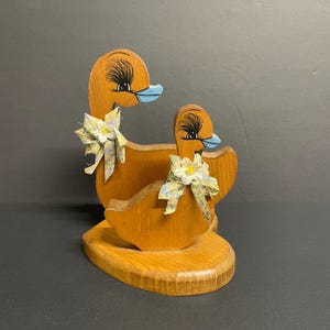 Könnte beinhalten: Holz-Serviettenhalter mit zwei Enten, eine größer als die andere. Die Enten sind braun bemalt mit blauen Schnäbeln und schwarzen Augen. Sie haben blaue und weiße Schleifen mit Blumen.