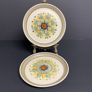 Op de afbeelding: Twee keramische borden met een bloemmotief. De borden hebben een wit midden met een grijze rand. Het bloemmotief heeft een centrale oranje cirkel omgeven door blauwe, gele en groene bloembladachtige vormen. De borden worden tentoongesteld op een decoratieve metalen standaard.