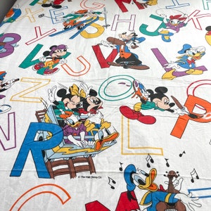 K&ouml;nnte beinhalten: Ein wei&szlig;er Baumwollstoff mit bunten Buchstaben und Disney-Figuren. Mickey Mouse, Minnie Mouse, Donald Duck und Goofy sind auf dem Stoff abgebildet. Der Text "The Walt Disney Co." ist unten in der Mitte des Stoffes gedruckt.