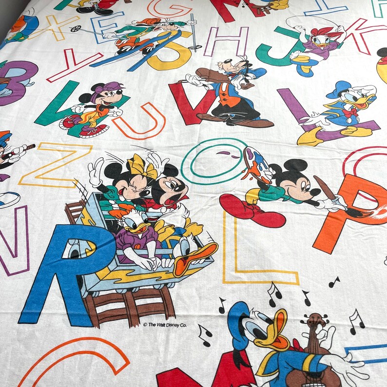K&ouml;nnte beinhalten: Ein wei&szlig;er Baumwollstoff mit bunten Buchstaben und Disney-Figuren. Mickey Mouse, Minnie Mouse, Donald Duck und Goofy sind auf dem Stoff abgebildet. Der Text "The Walt Disney Co." ist unten in der Mitte des Stoffes gedruckt.