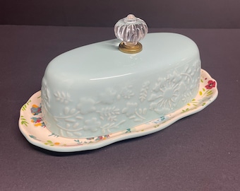 May include: A light blue ceramic butter dish with a floral design and a clear glass knob on the lid. The dish is sitting on a matching floral patterned plate.