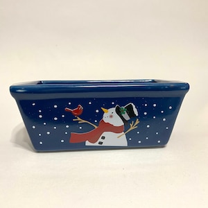 Puede incluir: Molde para pan de cerámica azul con un diseño de muñeco de nieve y cardenal. El molde tiene puntos blancos sobre un fondo azul.