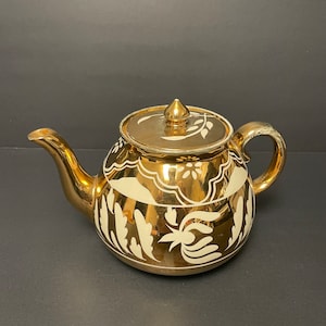 May include: A metallic gold teapot featuring a reflective finish and a white floral pattern. The teapot has a curved spout, a rounded handle, and a matching lid with a pointed finial. The white design includes leaf motifs and scalloped borders.