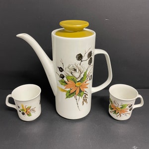 May include: A white ceramic coffee pot with a yellow lid and floral design. The pot is accompanied by two matching white ceramic mugs with floral designs.