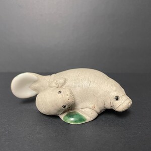 Op de afbeelding: Een keramische sculptuur van een zeekoe en kalf in een lichtgrijze kleur. De zeekoe heeft een afgerond lichaam met een kleine kop en donkere ogen. Het kalf ligt naast de zeekoe. De sculptuur heeft een groene accent op de basis.