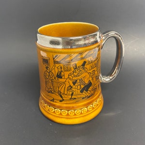 May include: A brown ceramic beer stein with a silver rim and a black and white illustration of people in a pub. The stein has a handle and a decorative pattern around the bottom.