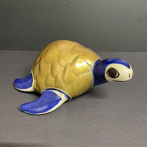 May include: A ceramic turtle figurine with a gold-coloured shell and blue flippers and head. The turtle's head is white with brown eye markings. The shell has a textured, scale-like pattern. A decorative item.