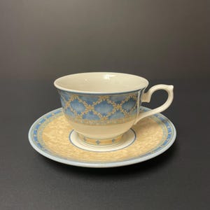 May include: A porcelain teacup and saucer set. The cup and saucer feature a blue and gold pattern with a cream-colored background. The cup has a curved handle and sits on a matching saucer.