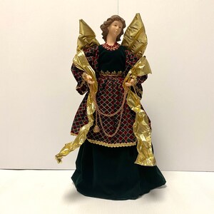 May include: A Christmas tree topper featuring an angel with gold wings and a red and black dress with gold trim. The angel is holding a string of gold beads.