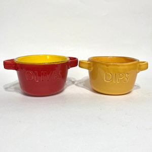 Peut inclure: Deux bols en céramique, un rouge avec l'intérieur jaune et le mot "OLIVES" sur le côté, et un jaune avec le mot "DIPS" sur le côté.