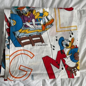 K&ouml;nnte beinhalten: Wei&szlig;er Stoff mit einem bunten Druck von Donald Duck und anderen Disney-Charakteren. Der Stoff zeigt einen roten, gelben und blauen Buchstaben "M" und einen gro&szlig;en orangen Buchstaben "G". Der Text "The Walt Disney Co." ist in Schwarz auf dem Stoff gedruckt.