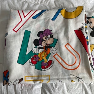 K&ouml;nnte beinhalten: Ein wei&szlig;es Baumwoll-Bettlaken mit einem bunten Druck von Mickey Mouse und anderen Comicfiguren. Das Laken zeigt eine Vielzahl von Buchstaben und Formen in Rot, Gelb, Blau, Gr&uuml;n und Orange.