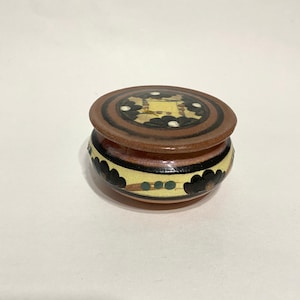 May include: A small, round ceramic trinket box with a lid. The box is decorated with a brown, black, white and yellow floral pattern.