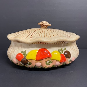 May include: A ceramic casserole dish with a lid, featuring a mushroom design. The dish is decorated with brown, yellow, red, and green mushrooms.