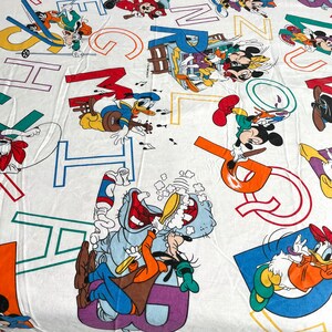 K&ouml;nnte beinhalten: Ein wei&szlig;er Stoff mit bunten Buchstaben und Comicfiguren. Die Figuren sind Mickey Mouse, Donald Duck, Goofy und Pluto. Die Buchstaben sind in verschiedenen Farben, darunter Rot, Blau, Gr&uuml;n, Gelb und Orange.