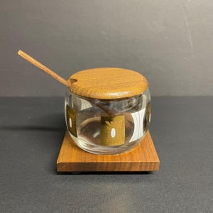 May include: A glass sugar dispenser with a wooden lid and a small wooden spoon. The dispenser has a geometric pattern in brown and white.