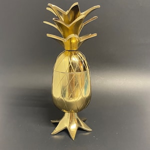 May include: A gold pineapple-shaped cocktail shaker with a detailed design. The shaker has a removable top and a base with three legs.