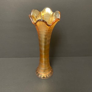 May include: A tall, amber-coloured iridescent glass vase with a flared, ruffled rim. The body of the vase features vertical ribbed detailing that extends from the base up to the neck, creating a textured appearance with a reflective, metallic sheen.