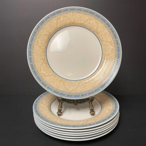 May include: A stack of white ceramic dinner plates with a beige and blue decorative border. The plates have a floral pattern on the rim and a blue border with a fleur-de-lis design. The plates are stacked on a decorative metal stand.