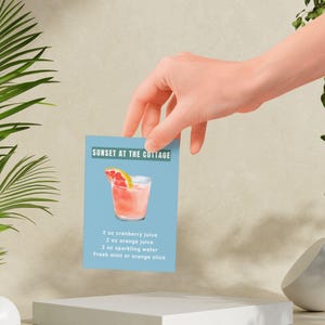 May include: A cocktail recipe card with a blue background and a pink drink with a grapefruit slice. The recipe is called "Sunset at the Cottage" and includes 2 ounces of cranberry juice, 2 ounces of orange juice, 2 ounces of sparkling water, and fresh mint or an orange slice.