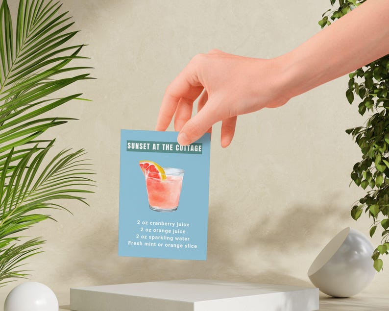 May include: A cocktail recipe card with a blue background and a pink drink with a grapefruit slice. The recipe is called "Sunset at the Cottage" and includes 2 ounces of cranberry juice, 2 ounces of orange juice, 2 ounces of sparkling water, and fresh mint or an orange slice.