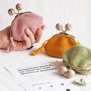 K&ouml;nnte beinhalten: Drei geh&auml;kelte Geldbeutel mit Metallverschl&uuml;ssen und Holz-Kn&ouml;pfen. Die Geldbeutel sind rosa, gelb und gr&uuml;n. Die Geldbeutel liegen auf einer wei&szlig;en Oberfl&auml;che mit einem Blatt Papier mit dem Text "Monedero B&aacute;sico de Crochet con Boquilla Redonda" auf Spanisch.