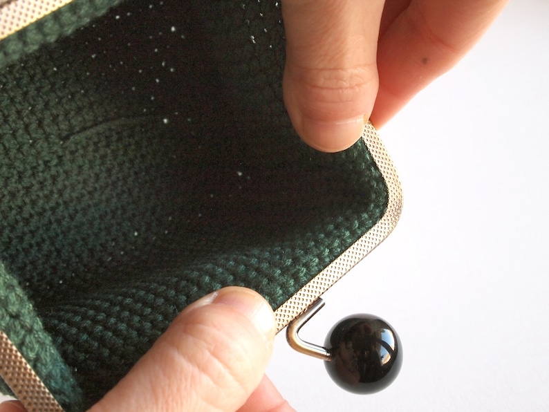 May include: A green crocheted coin purse with a silver metal frame and a black button closure.