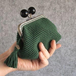 May include: A green crocheted coin purse with a silver and white trim. The purse has a tassel and two black round clasps.