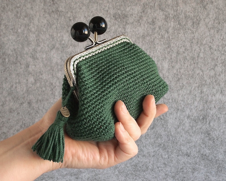 May include: A green crocheted coin purse with a silver and white trim. The purse has a tassel and two black round clasps.