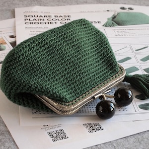 May include: A green crocheted coin purse with a metal clasp and a tassel. The purse is lying on a white paper with a QR code and the text "the https://youtu.be/JUC4ETPA".