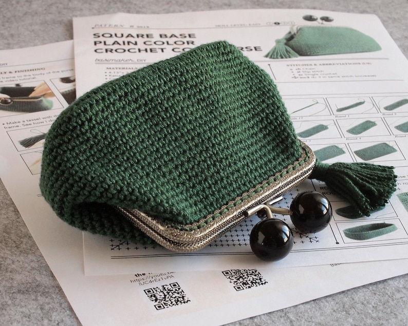 May include: A green crocheted coin purse with a metal clasp and a tassel. The purse is lying on a white paper with a QR code and the text "the https://youtu.be/JUC4ETPA".