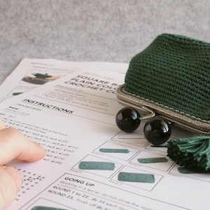 May include: Instructions for crocheting a square, green crochet coin purse with a silver clasp and black beads. The instructions include a base and going up sections. The purse is shown in the image.