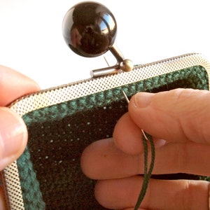 May include: Close-up of a hand sewing green fabric to a silver metal frame. The frame has a black metal knob on top.