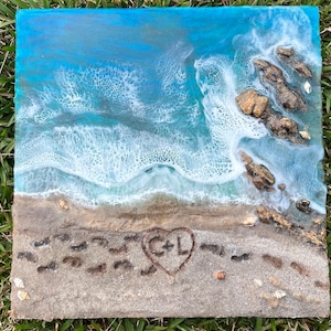 May include: Square resin art piece depicting a beach scene. The artwork features a blue ocean with white waves crashing on a sandy shore. Brown rocks and seashells are visible. The sand has footprints and a heart with the initials "C+L".