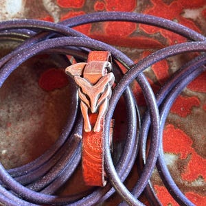 May include: A leather bracelet with a silver fox head clasp. The bracelet is a reddish-brown color with a paisley pattern. The bracelet is surrounded by a coiled, dark purple cord with a glittery finish. The background is a red and brown speckled surface.