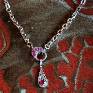 May include: A silver chain necklace with a teardrop-shaped pendant featuring a pink gemstone. The pendant is attached to a silver toggle clasp with three pink gemstones.