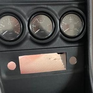 Puede incluir: Panel de salpicadero negro con tres indicadores redondos, un recorte rectangular y dos agujeros circulares. Los indicadores tienen agujas blancas y caras negras con marcas blancas. Los indicadores marcan "12 Volt", "2 RPM" y "80 PSI".